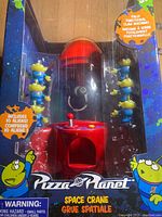 Front view of boxed Pizza Planet Space Crane with 5 green alien toys visible and text on box.