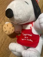 Close-up of Snoopy plush face, apron with embroidered text 'Happiness is a Warm Cookie' and plush cookie on paw.