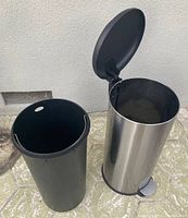 Tall stainless steel kitchen trash can with black plastic removable liner next to it on a carpeted floor outside.
