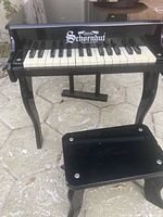 Full view of black Schoenhut toddler piano with keyboard and matching black toddler piano stool on outdoor stone surface.