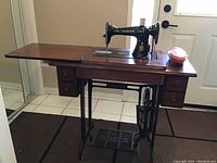 Front view of the black Singer sewing machine installed in the wooden treadle cabinet with drawers and fold-out top.