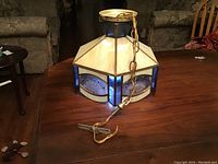 Beige and blue leaded stained glass light fixture with hexagonal shape placed on wooden surface. Shows the fixture and hanging chain with electrical cord.