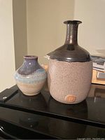 Front view showing the two vases side by side on a black surface with a beige wall background. Taller vase shows the smooth metallic bronze glaze on the neck and the sand-like textured bottom with a round shape. Smaller vase shows layered glaze colors.