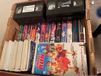 Photo showing a collection of VHS tapes in original cases, including Chicken Run and others, stacked in a cardboard box.