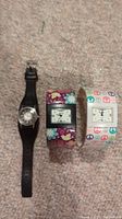 All three watches displayed side by side on carpet. One black leather band Roots watch and two wide plastic band Suisse watches with colorful floral and peace symbol patterns.