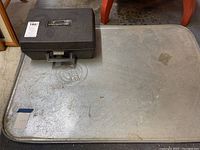 Photo of metal oil catcher plate with black battery booster cables case on top, showing surface wear and dirt.