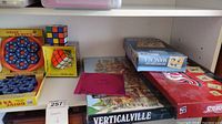 Photo shows a shelf with multiple board games and puzzles including Scrabble, Mancala, Hoyle Board Games, Verticalville, Rubik's Cubes, and two Drive Ya Nuts puzzles.