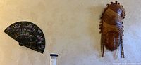 Photo showing both the dark floral oriental hand fan and the light-toned ethnic wood carved wall mask hung against a beige textured wall.