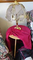 Lamp with gold metal frame and six glass panels placed on a round light wood table covered with a red tablecloth, showing side view including part of surrounding furniture.