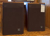 Pair of JBL speakers side by side on wooden surface under window.