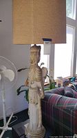 Side view of a tall pottery figure lamp with beige fabric shade, showing carved details of robe and figure.