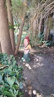 Gnome statue with red hat, yellow shirt, blue pants, and green base seen from front and side in garden setting under trees.