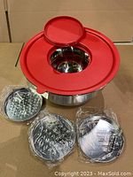 Front view of the stainless steel mixing bowl with red lid featuring a central opening. Three stainless steel discs for grating, shredding, and slicing are displayed next to the bowl, wrapped in protective plastic.
