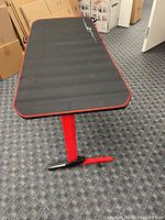 Oblique view showing length and design details of gaming desk with black surface, red trim edges, and red T-leg supports on blue patterned carpet.