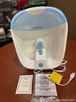 Homedics Deep Soak Foot Spa front view showing the white and blue spa basin with massage rollers and control switches, power cord coiled below, accompanied by paperwork and trial salt packet.