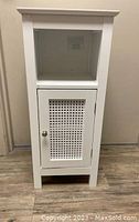 Front view of white bathroom storage cabinet showing open shelf on top and rattan style door on bottom with knob.