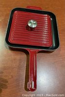 Square red cast iron grill pan with matching lid set on wood surface, showing full view including handle and lid knob.