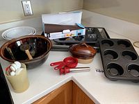 Photo showing all items in the lot including a brown pottery dish with lid, measuring spoons, dough cutter, ceramic holder with spoons, and baking pans.