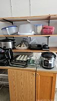 Overview of shelving with several baking sheets, a muffin pan, plastic containers, a white cake carrier, rice cooker, and crock pot on kitchen shelving.