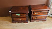 Pair of solid wooden bedside tables side by side on a wood floor against a brown wall, showing the fronts and tops of both tables with drawer handles visible.
