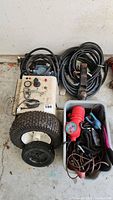 Photo showing several electrical cords, a sump pump, a container with tie down straps, and two wheels with tires.
