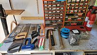 Photo of hand tools including saws, hand hammers, screwdriver, and staple gun on a workshop table.