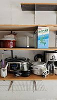 Wide view of multiple kitchen items on wooden shelves including slow cooker, crock pot, coffee carafe, frying pans, and storage containers.
