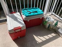 Two coolers side by side, red and white Igloo cooler in front, red and white Coleman cooler with turquoise lid behind, plus assorted garden tools and spray bottles on the side.