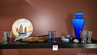 Full display showing the electric blue vase, decorative plate with ship, multiple colored glass goblets, wooden tray, and glass paperweights on a black shelf.