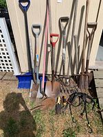 Photo showing full assortment of garden tools, including snow shovel, shovels, pitchfork, hoe, hand tools and bungee cords arranged against shed wall outdoors in grass.