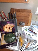 Front view showing green covered baked dip dish in box next to knife block, cutting board, and stacked Corelle plates with utensils.