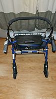 Front view of blue mobility walker showing padded handles with brake levers, foldable black seat/storage basket, and caster wheels on wooden floor.