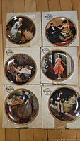 Six Norman Rockwell collector plates placed on their boxes, showing detailed artwork of various scenes including laundry, a dining couple, writing woman, Christmas girl, sewing woman, and ironing man.