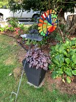 Tall black wicker planter outdoors with purple/burgundy foliage plant inside and colorful garden pinwheel behind it.