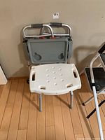 Photo of white plastic shower bench and folded gray walker leaning against wall with bed assist rail handles and stepping stool partially visible.