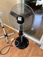 Black pedestal fan with three black blades and solid black base, standing on wood floor near glass door.