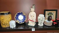 Shelf display of various Asian inspired decorative items including statues, dishes, jars, framed calligraphy, and tea cups