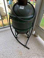 Front view of green ceramic kamado grill mounted on metal cart with casters. Hose draped over front and sticker 162 on lid.