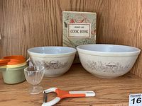 Photo showing two Pyrex bowls with Forest Fancies brown mushroom pattern, vintage cook book, measuring cups, egg cup, and vegetable peeler arranged on wooden surface.