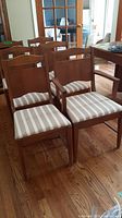 Overall view of six wooden dining chairs, one with armrests, upholstered with striped beige and white fabric seat cushions.