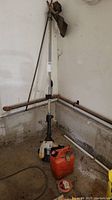 Full view of Ryobi gas powered trimmer with straight shaft leaning against wall, red fuel can, and spool of extra trimmer line on floor.