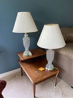 Pair of matching table lamps on wooden nesting tables against a blue wall, both lamps have clear patterned glass bases and white textured fabric shades.