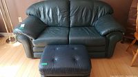 Front view of green leather love seat with matching ottoman on light wood floor.