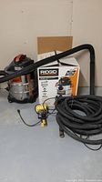 Wide shot of the Ridgid wet/dry vac showing the stainless steel tank, flexible hose, yellow electrical device, and the cardboard box behind the unit.