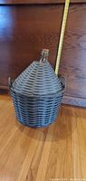 Full view of light green glass carboy with gray woven wicker basket cover and handles, showing size with measuring tape.