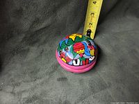 Top view of the closed trinket dish with colorful folk art designs on the lid and base. The dish is shown next to a measuring tape indicating approximately 2.5 inches in diameter.