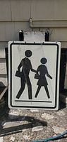 Front view of rectangular pedestrian crosswalk sign with adult and child silhouettes, metal brackets and hanging chains visible at top