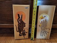 Front view of two animal carvings with measuring tape showing height about 9-10 inches. One depicts a howling black wolf with a raccoon and sun, the other a bald eagle. Both signed Billy Mays.