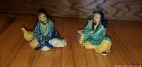 Lot of two vintage Asian-style ceramic figurines on wooden surface, one holding a gourd and wearing blue and green robes, the other with blue hood and yellow pants sitting on a small box.