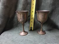 Pair of vintage brass goblet cups approximately 7 inches tall shown with a measuring tape for scale. Patina and wear visible on the surface.